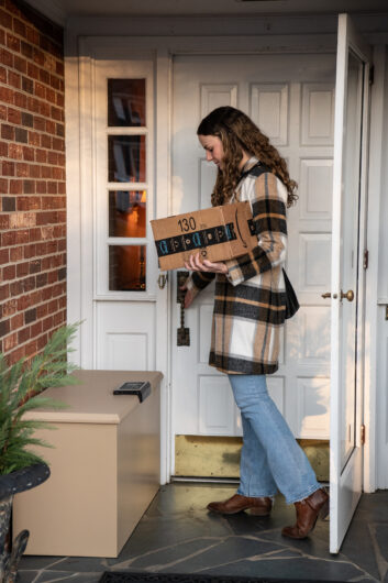 A woman retrieves a cardboard box from a steel parcel protection box at her front door. 