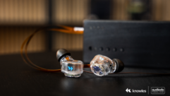 A pair of wired IEMs with the Knowles logo rest on a dark tabletop. In the background, they are connected to a DAC/amp system. The right bottom corner features the Knowles and AudioDo logos.